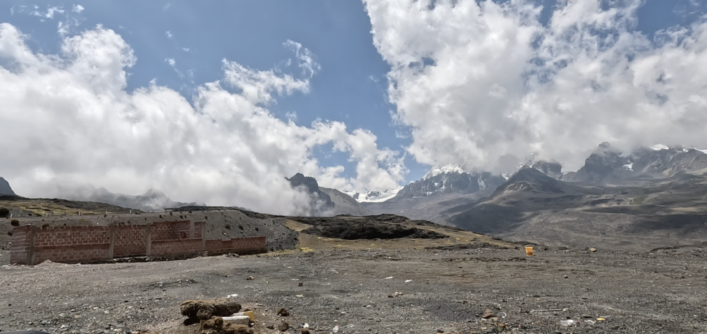 The high mountain pass at Abra Pirhuayani in Peru, showing a half-built brick shack in the foreground and the snow-capped Ausangate peaks behind clouds.
