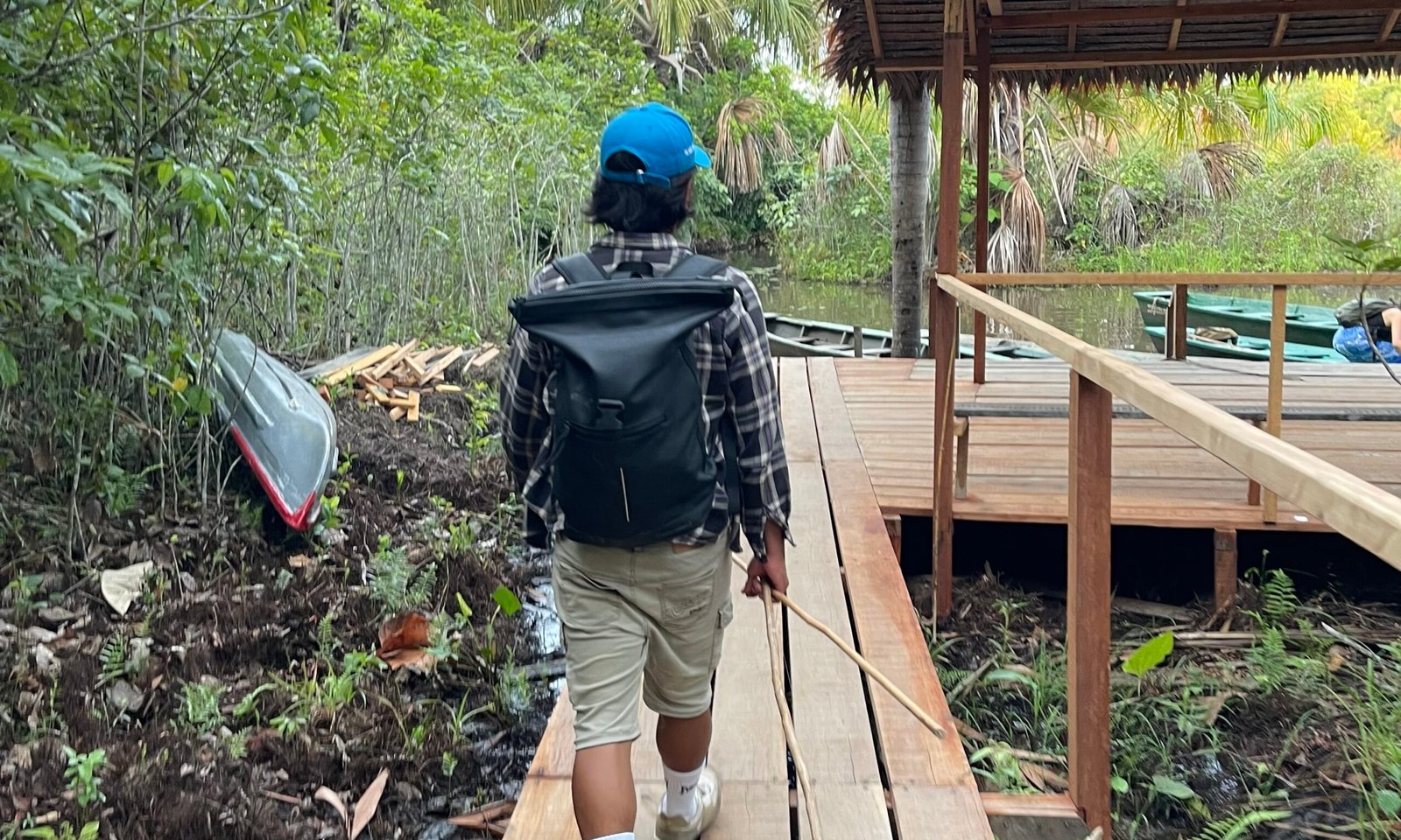 Marco, a local guide, carrying a dry bag and traditional bamboo fishing poles while walking toward a canoe dock in the Peruvian Amazon rainforest.