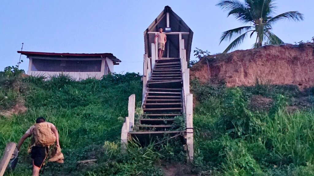 A man carrying heavy luggage up a steep, muddy riverbank toward a lodge in the jungle.