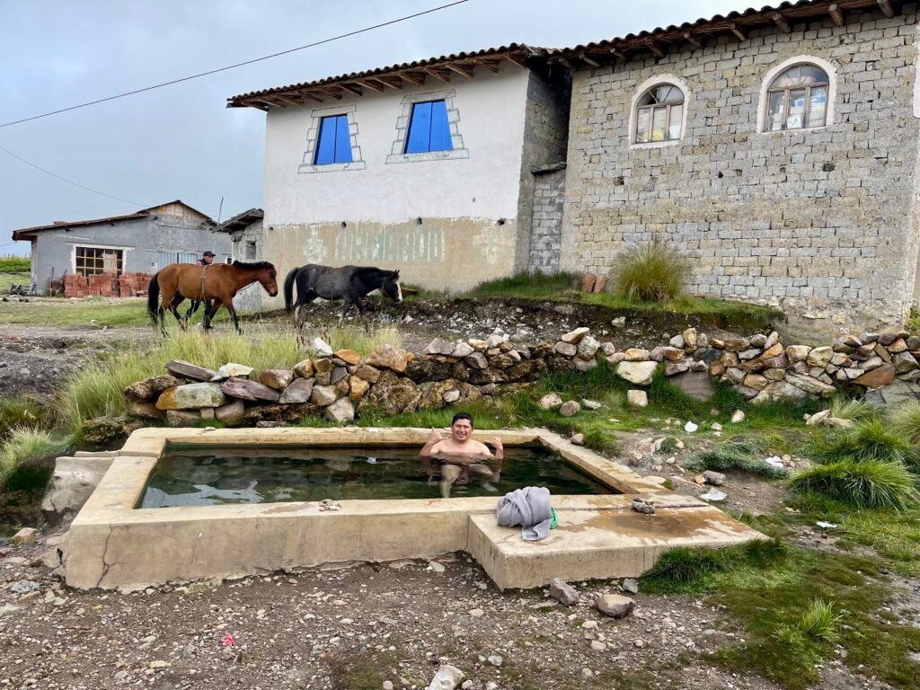 A traveler relaxing in the outdoor medicinal hot springs of Pacchanta village with the snow-capped peaks of Apu Ausangate visible in the distance.