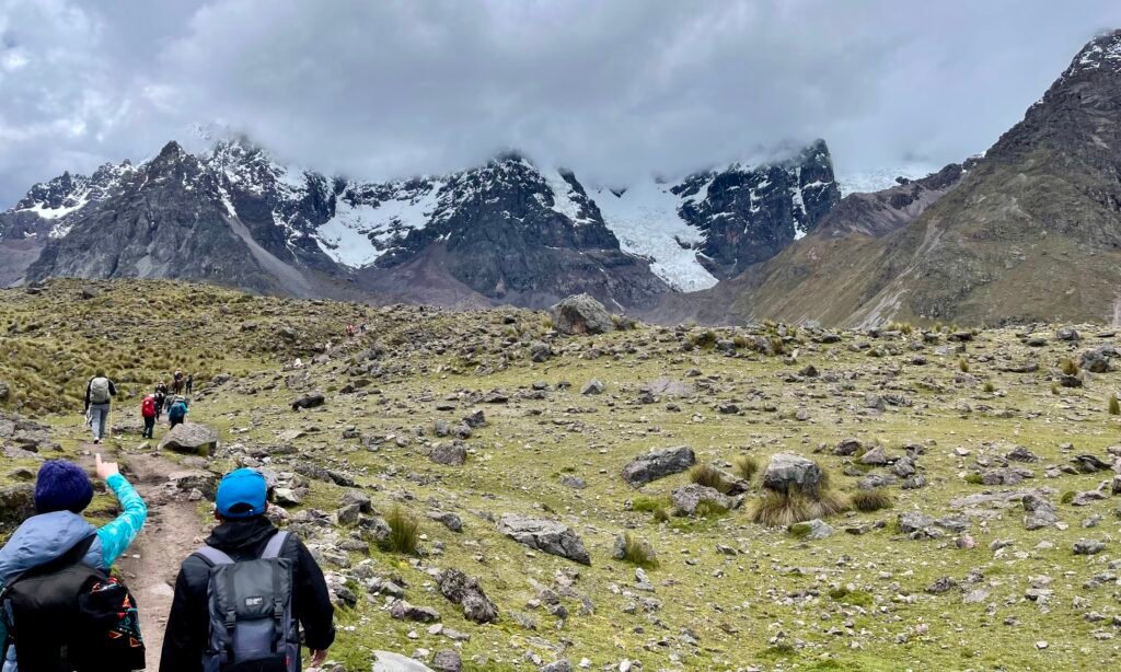 A vast Andean valley with steep green slopes and distant hikers, showing the massive scale of the ascent toward Ausangate.