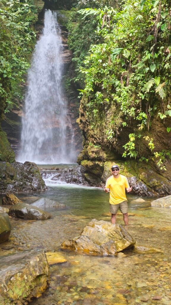 A traveler in a yellow shirt stands in a shallow river at the base of a tall waterfall in the Camanti Cloud Forest, Peru, on the drive between Cusco and Puerto Maldonado.