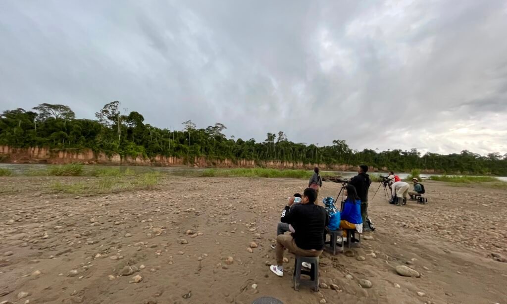 A group of travelers sitting on small plastic stools out in the open on a wide sandbar in the middle of the river, waiting over a hundred meters away from the empty Colpa Chuncho clay cliff face under a gray, overcast sky.
