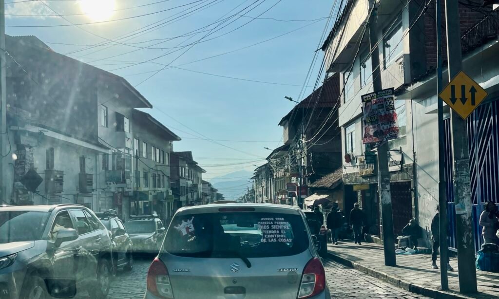 A gritty urban street in Cusco, Peru, at the start of the drive to Puerto Maldonado. The photo shows tangled overhead power lines, local traffic, and the grey, industrial atmosphere of the city's outskirts.