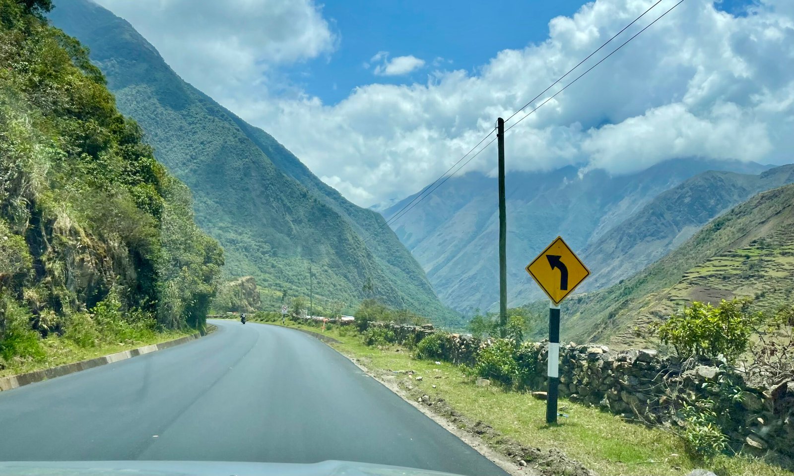 A driver's perspective from inside a car on the 30C Interoceanic Highway in Peru. The view shows a paved road curving past ancient agricultural terraces on a steep hillside, with green Andean mountains rising into a heavy cloud layer.