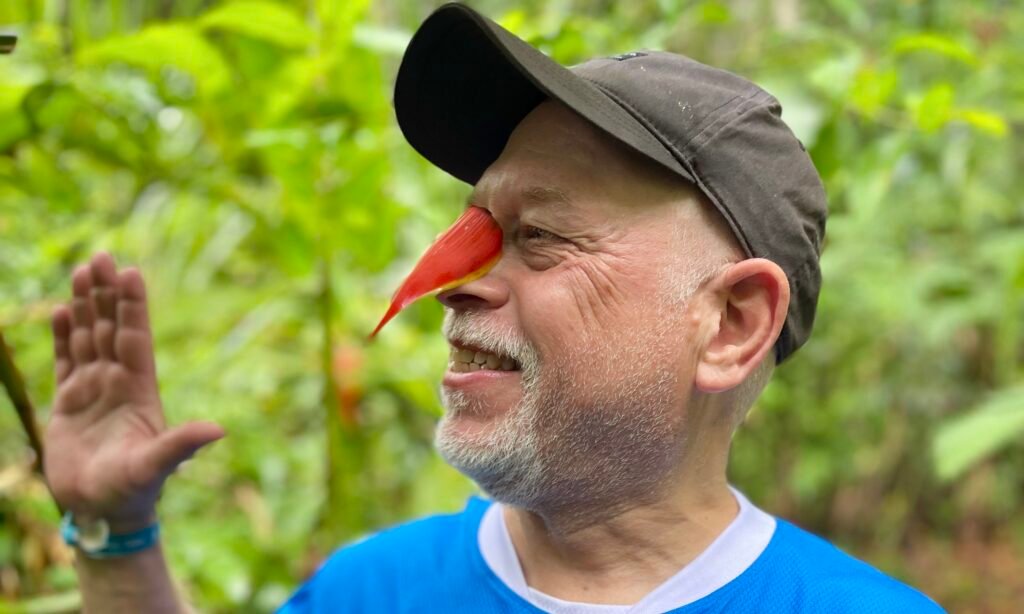 Dave smiling with a bright red wild flower balanced on the bridge of his nose while on a jungle trail.