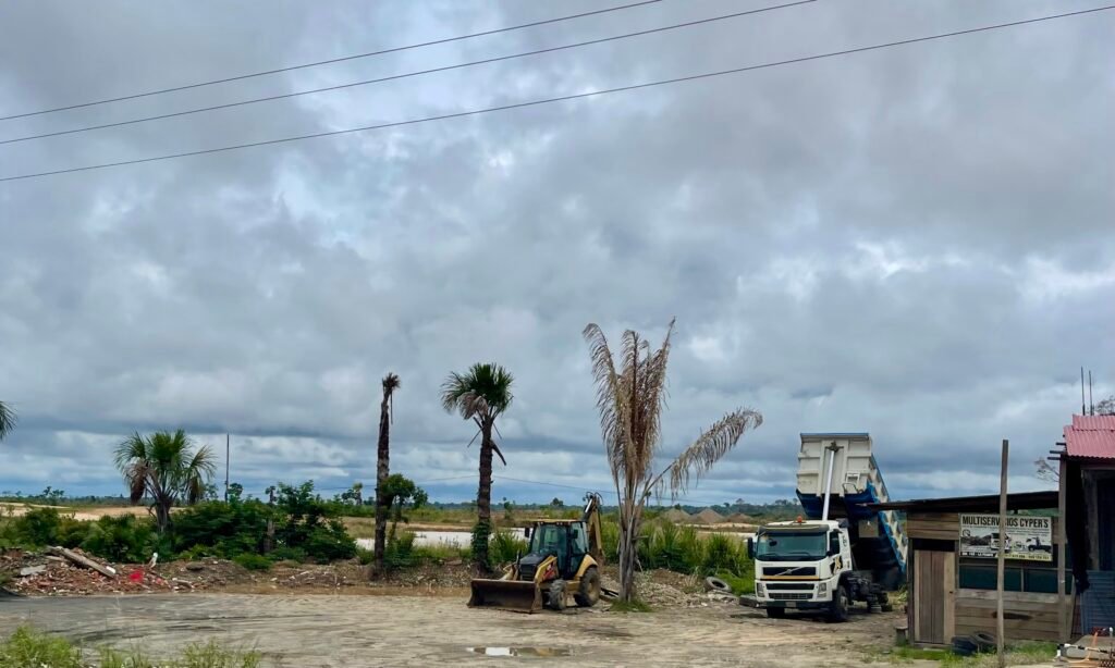 A gritty, close-up crop of La Pampa illegal mining, showing a large yellow bulldozer on the left, a corrugated metal shanty shack on the right, and a solitary dead palm tree in the center of an expanse of orange sand.