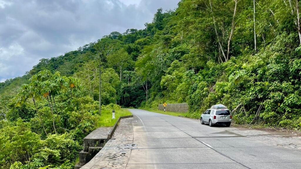 A silver Honda CR-V parked on the shoulder of the 30C Interoceanic Highway, surrounded by a dense, towering wall of lush green tropical jungle under a cloudy sky.