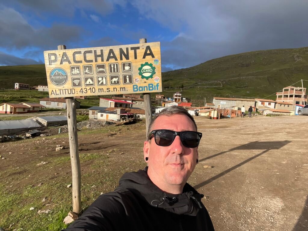 A wooden welcome sign for the village of Pacchanta, Peru, showing an altitude of 4,310 meters above sea level with a background of Andean mountains.