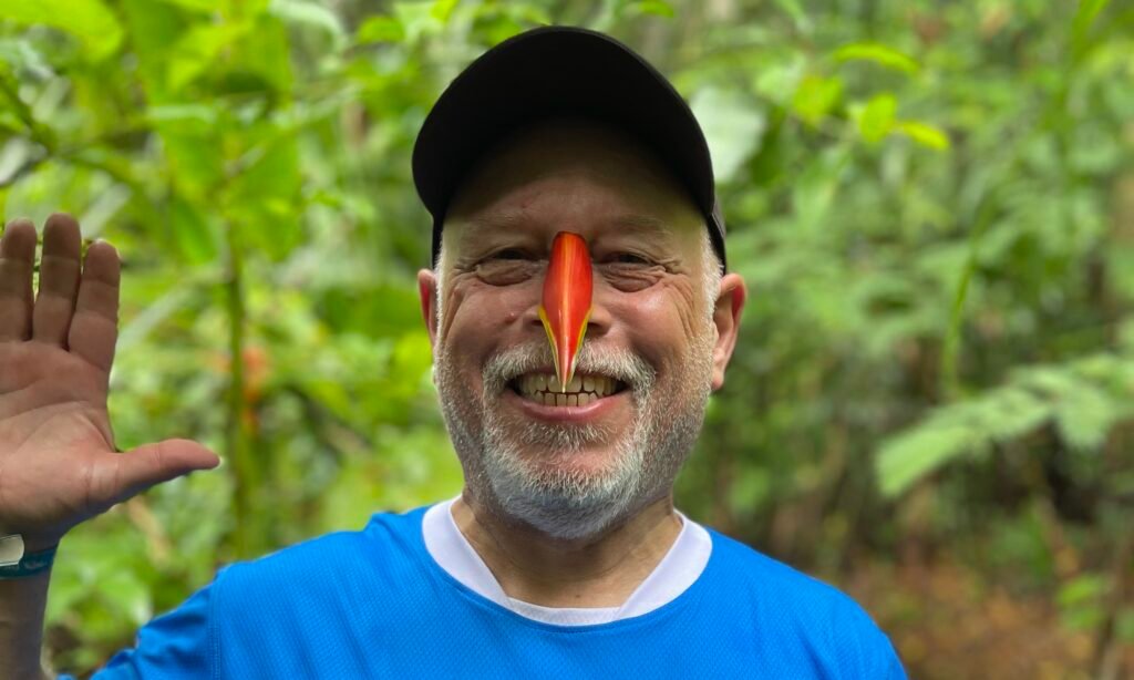 A close-up of a traveler smiling on a jungle trail at Monkey Island near Puerto Maldonado, wearing a split bird of paradise flower petal over his nose to look like a parrot beak during a guided tour.