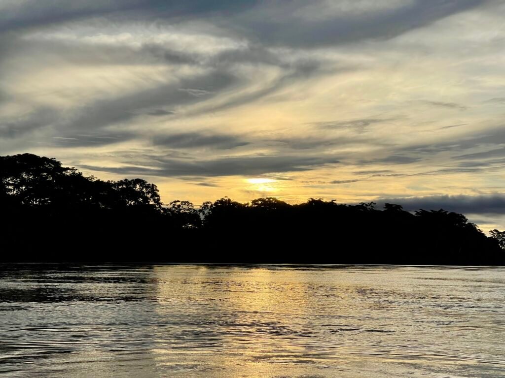 A wide, cinematic sunset over the Tambopata River in the Peruvian Amazon. The sky is a gradient of deep orange and purple, reflecting in the calm river water with the dark silhouette of the jungle canopy on the horizon.
