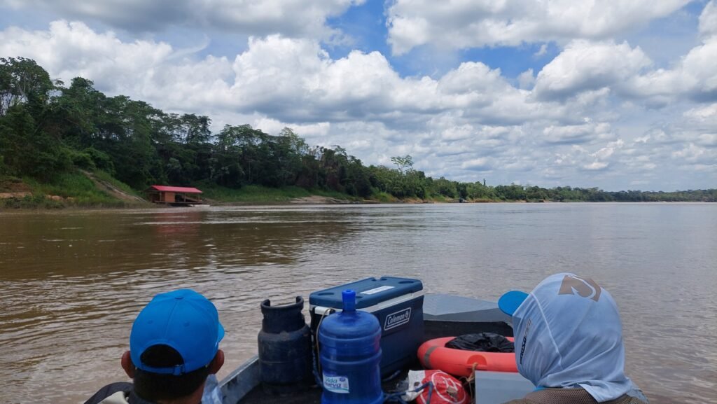 View from the front of a small motorboat traveling down the Madre de Dios River, showing a cooler, a gas canister, and a water bottle stowed at the bow.