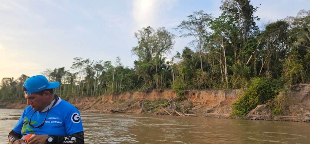A view from a boat on a wide brown river in the Peruvian Amazon, with local guide Percy on the left and a steep clay bank with collapsing trees on the right.