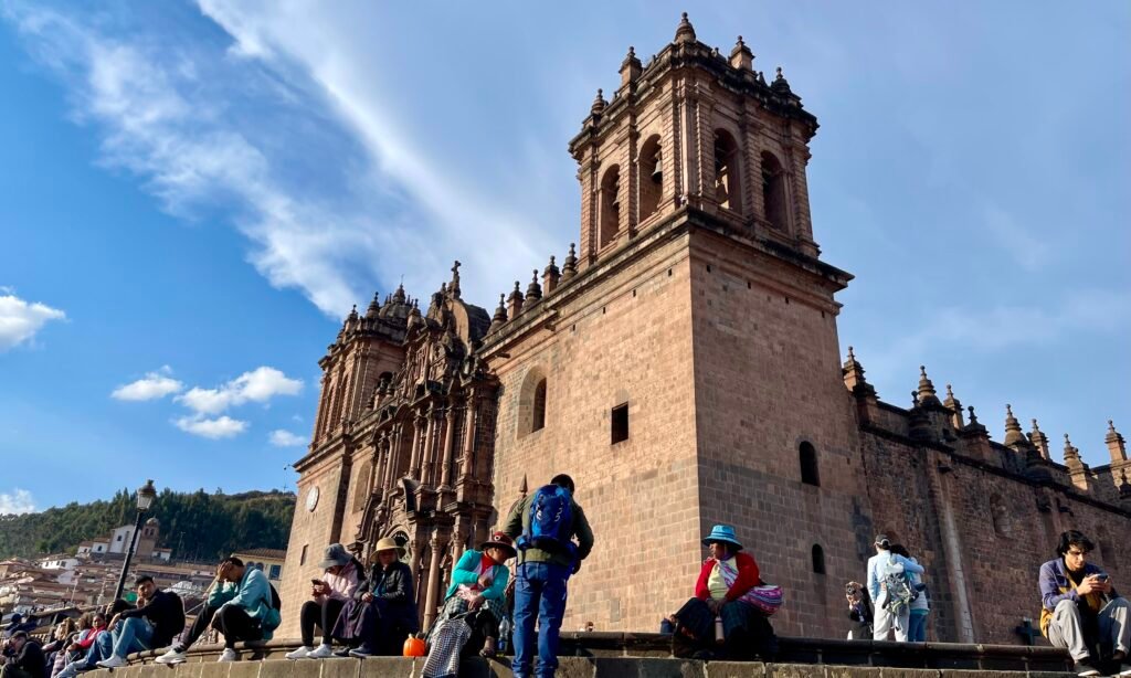 Ground-level view looking up at the Cusco Cathedral from the steps, featuring local Andean women in traditional dress amidst a crowd of tourists taking selfies.