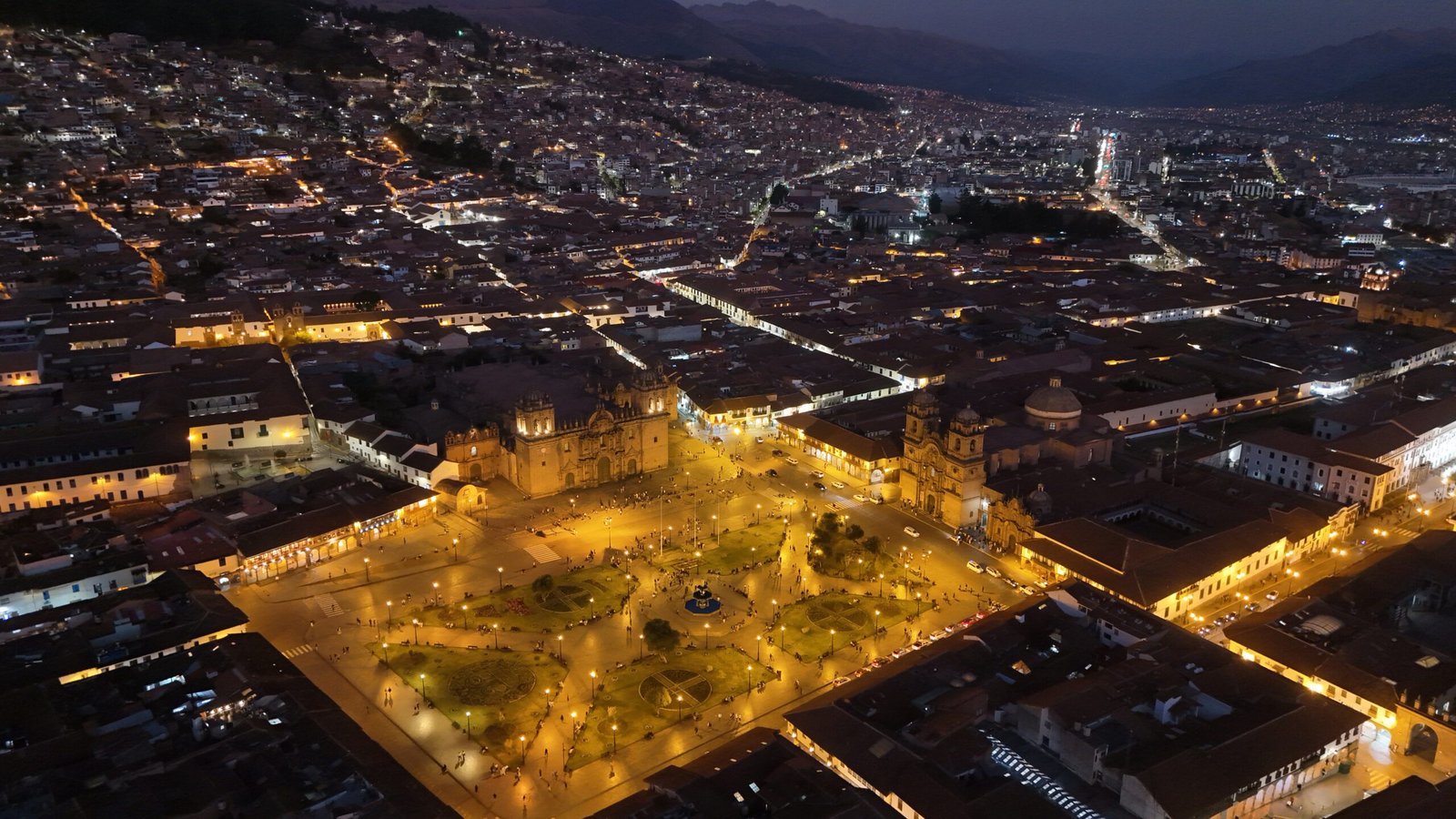 Aerial view of Cusco's Plaza de Armas illuminated at night, looking down into the valley as the city lights stretch into the distance.
