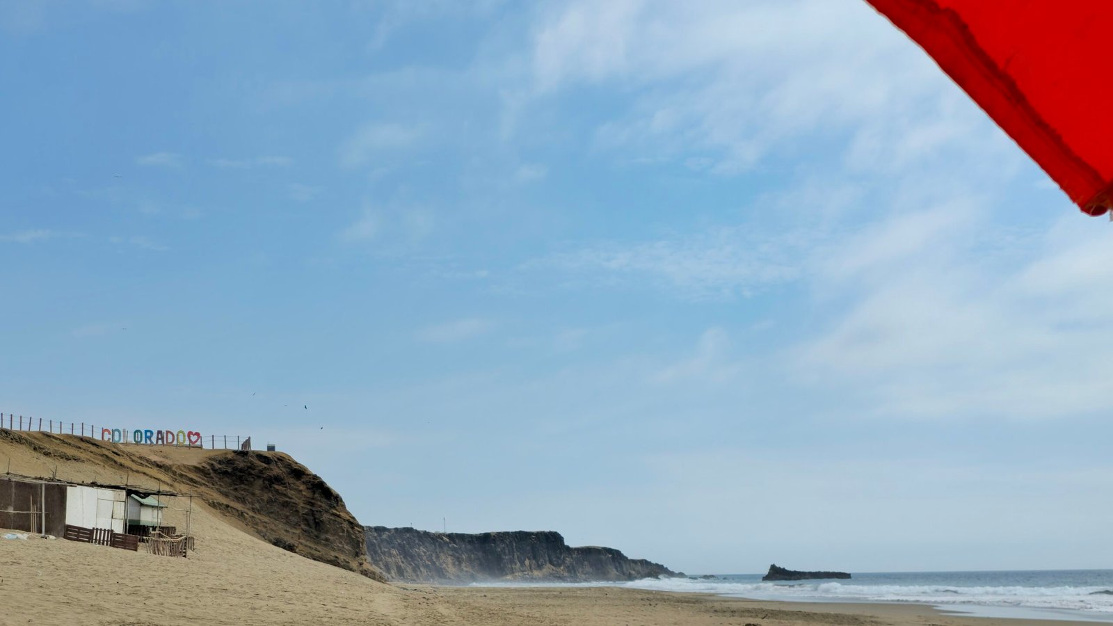 A bright red beach umbrella framing a view of Playa Colorado in Huacho, Peru, with sandy dunes and coastal cliffs in the background.