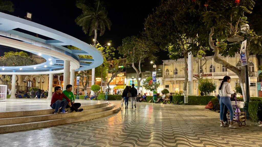 Locals relaxing in the Plaza de Armas in Huacho, Peru, at night, with a street vendor selling coffee and hot chocolate to the right.