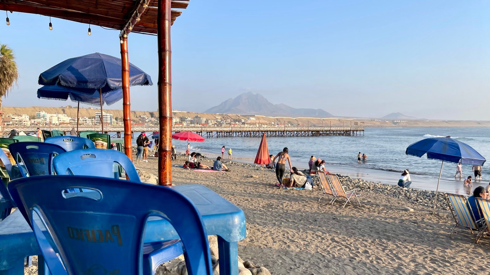 View from a local beach bar on Playa Varadero in Huanchaco, Peru, looking toward the pier and mountain coastline.