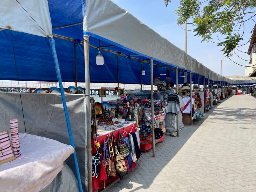 A row of covered artisan market stalls in Huanchaco displaying traditional Peruvian handicrafts, textiles, and pottery under a blue canopy.