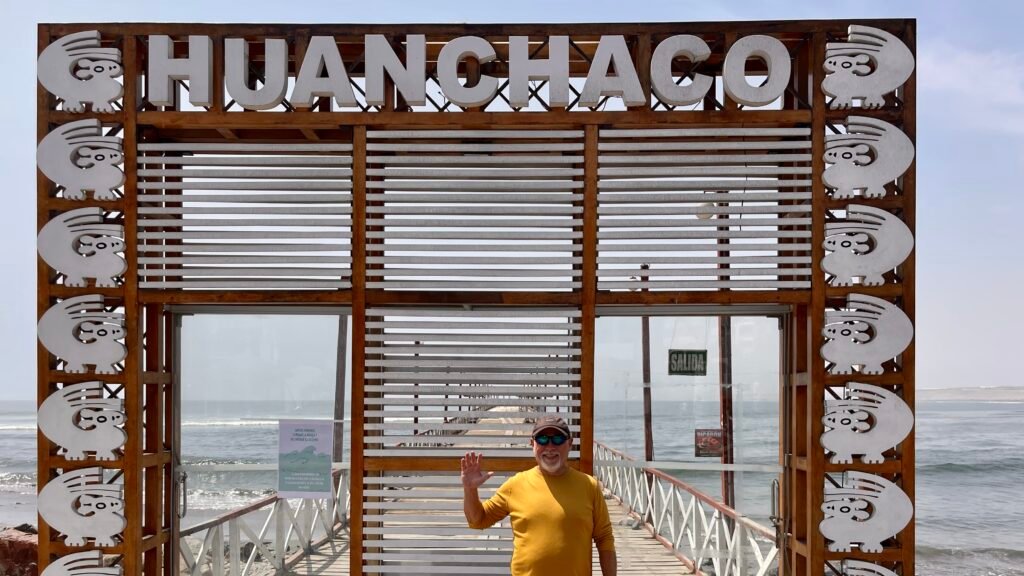 Dave waving in front of the iconic wooden entrance archway to the Huanchaco pier, featuring traditional Moche-style artwork on the frame.