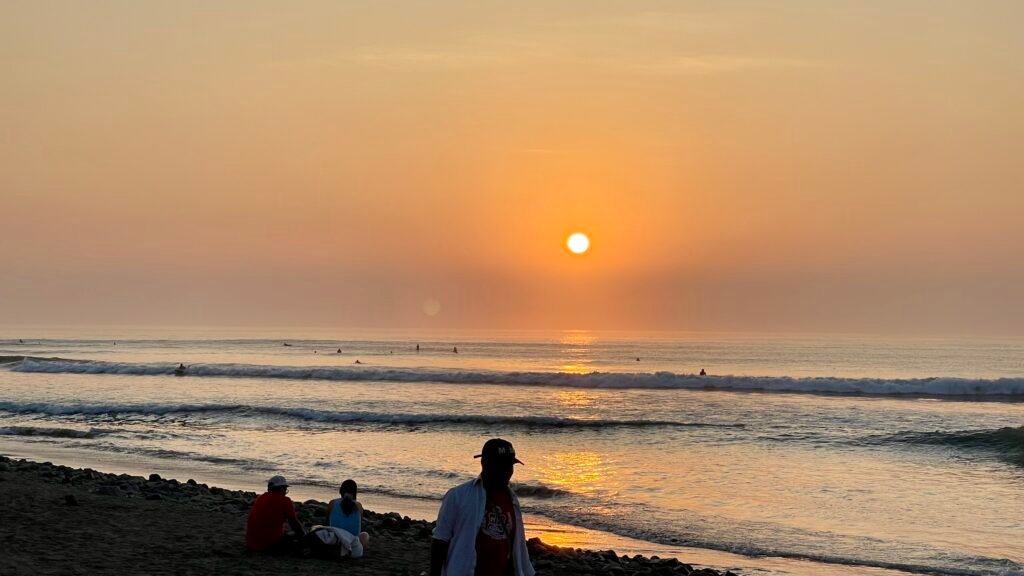 A golden hour sunset over the Pacific Ocean in Huanchaco, with surfers waiting for waves and people relaxing on the beach.