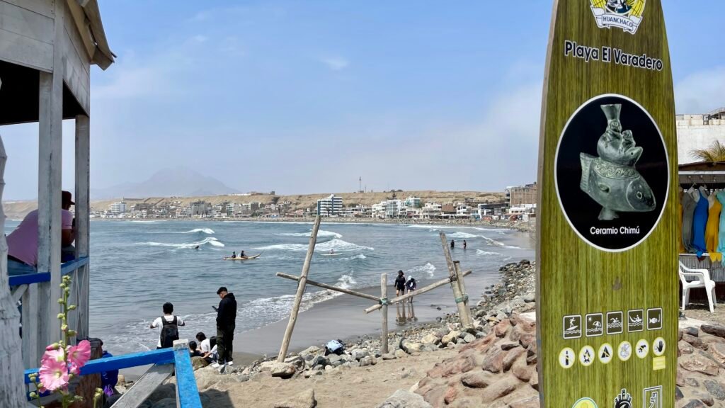 Informational surfboard sign at Playa El Varadero in Huanchaco, Peru, overlooking surfers and caballitos de totora in the water.