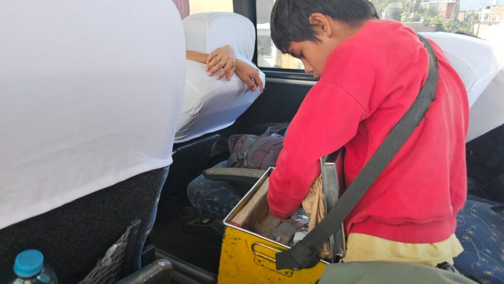 A young boy in a red sweater selling snacks and drinks from a metal box to passengers on a bus departing Huarmey, Peru.
