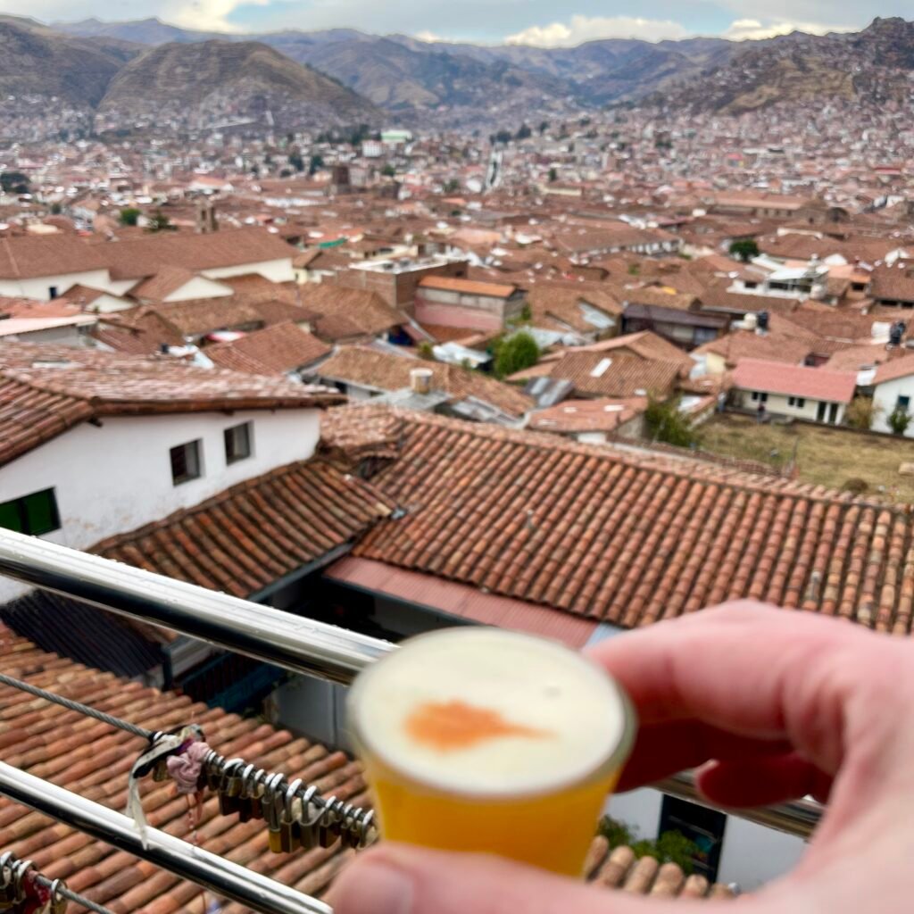 A glass of Pisco Sour held against a panoramic backdrop of Cusco’s terracotta rooftops and Andean mountains.