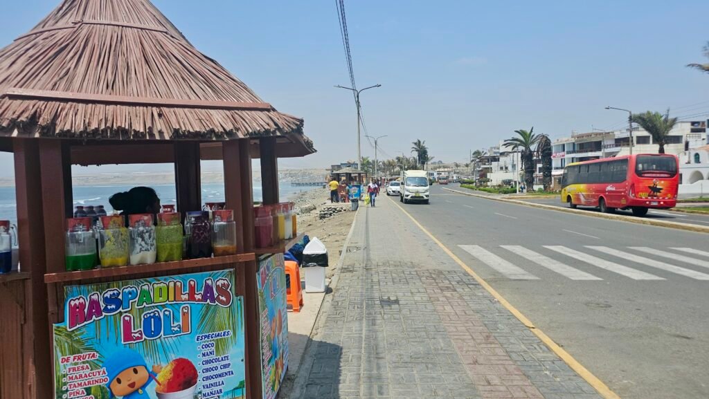 A roadside view in Punta Huanchaco, Trujillo, showing a local fruit juice and shave ice stand overlooking the Pacific Ocean with a bus passing by.