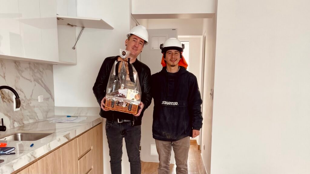 Kieran Proctor and his contractor, both wearing white hard hats, stand in a modern, newly finished kitchen in Lima. Kieran, on the left, holds a gift basket, while his contractor stands next to him smiling.
